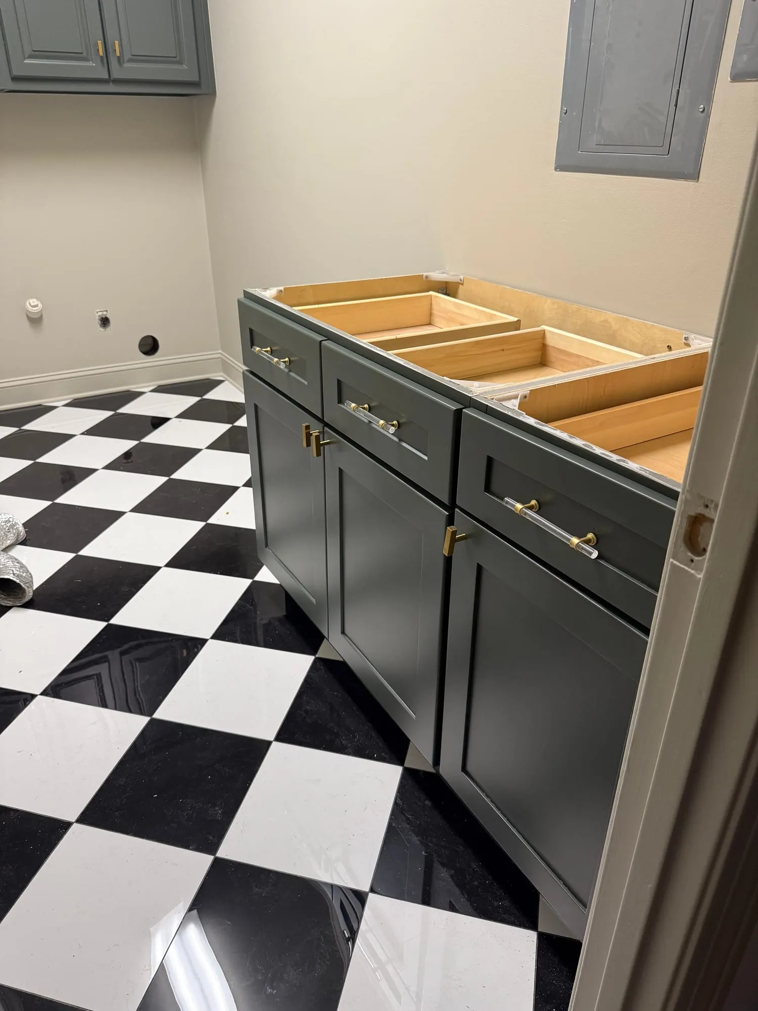 A bathroom base cabinet refinished in soft slate green with shaker doors and brass-and-glass pulls, drawer boxes pulled out mid-install, over a black-and-white checkerboard floor.