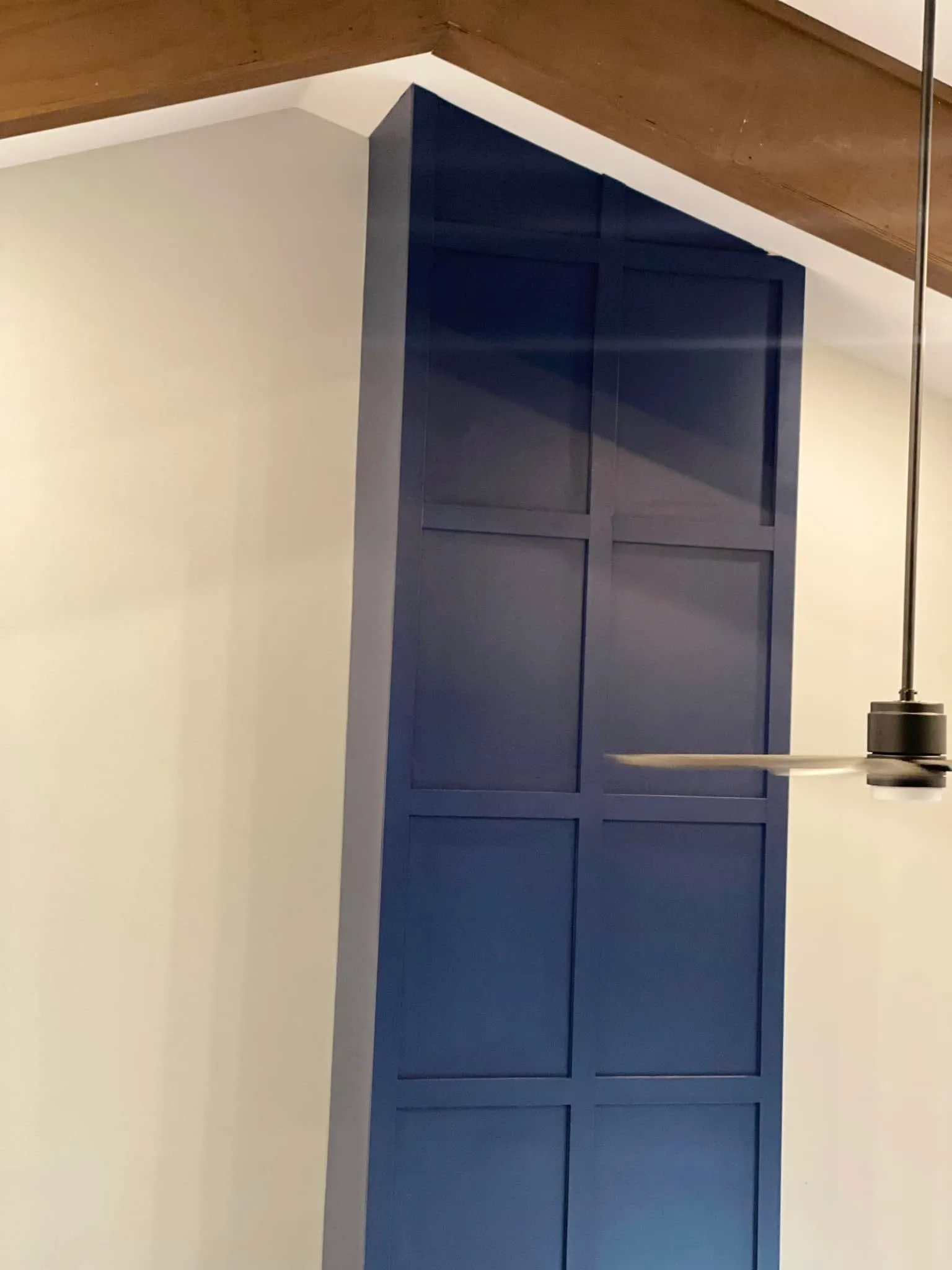 A tall built-in cabinet painted deep navy with shaker panel detail, set against white walls under a stained wood beam.