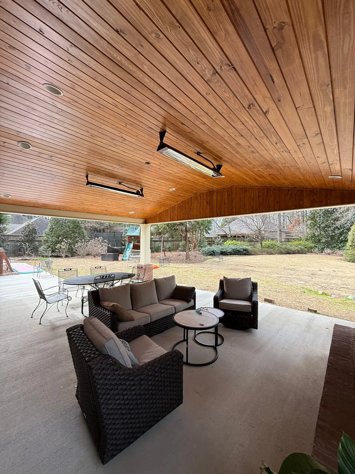 A sloped patio ceiling clad in honey-stained wood planks above outdoor seating, looking out across a wide back yard.
